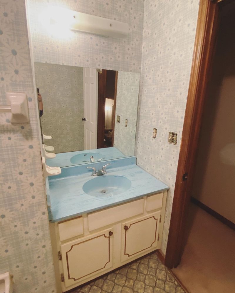 Close-up of original vanity with blue laminate top, wallpaper, and dated light fixture
