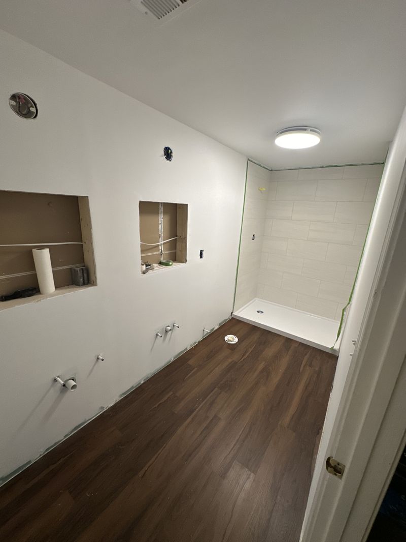 Bathroom renovation in progress with dark wide-plank LVP flooring installed, shower tile started, and vanity plumbing stubbed out
